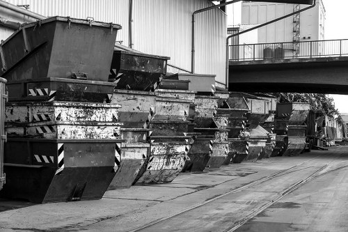 A row of large, heavy-duty metal skips filled with mixed waste materials, positioned along an industrial asphalt surface adjacent to a warehouse or factory building. The skips are constructed from dark, weathered steel with visible rust patches, scratches, and graffiti stickers, indicating extensive use. They are arranged side by side, with some slightly leaning or overlapping, and their open tops revealing a mixture of debris and refuse inside. In the background, a section of the building wall, made of corrugated metal panels, can be seen along with a raised walkway or balcony with metal railings. The area around the skips appears utilitarian, with environmental elements typical of a waste management or rubbish collection site. The black-and-white photograph emphasizes the textures of the metal surfaces and the industrial setting, conveying a scene related to commercial rubbish removal services in Marylebone.