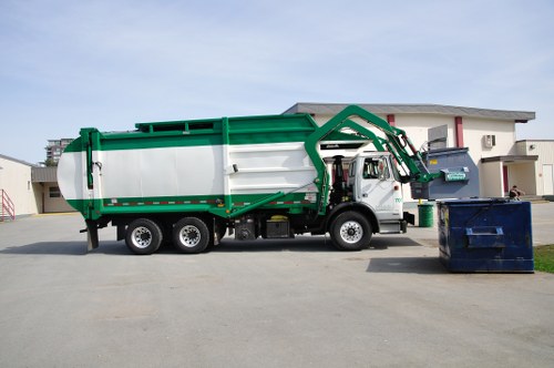 A large commercial waste collection truck parked on a paved driveway in a residential area, with a white cab and a green and white waste container. The container has a smooth metal surface with horizontal ridges, and the truck’s mechanical arm is in a folded position alongside it. To the right of the truck, there is a blue recycling bin and a smaller black waste bin placed on the ground. In the background, a modern two-storey white house with a flat roof and large windows is visible, with minimal landscaping and a lawn area. The sky is partly cloudy, and the scene is evenly lit, indicating daytime. This image relates to rubbish removal services provided by Commercial Waste Marylebone, highlighting waste collection operations in an urban neighbourhood within the Marylebone district, close to central London postcode area.