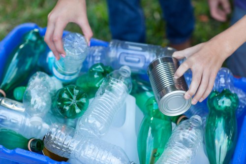 A person’s hands are visible placing a clear plastic water bottle into a blue bin filled with various discarded plastic bottles, some green and others transparent, during a waste collection or recycling effort. The bottles vary in size and shape, and are made of smooth, hard plastic, with some displaying ridged caps while others are open. The person's arms are shown from the forearms downward, wearing casual clothing, and the scene appears outdoors with a grassy background that suggests a garden or park environment. The image context relates to rubbish disposal and recycling practices, consistent with commercial waste management services in Marylebone, as offered by Commercial Waste Marylebone. The focus is on the sorting and handling of plastic waste, emphasizing environmentally conscious waste separation at the point of collection or disposal.