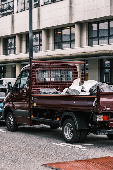 A small burgundy flatbed truck parked on a city street in front of a modern office building with large glass windows and concrete panels. The truck's cargo bed is filled with a variety of waste materi