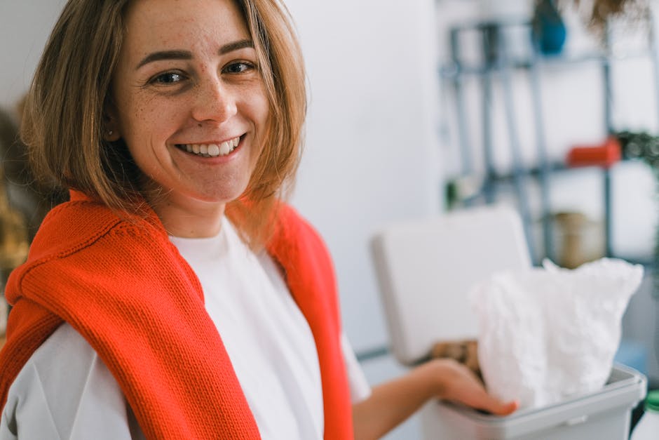 A woman with shoulder-length light brown hair and fair skin, smiling at the camera, is dressed in a white shirt with an orange knitted cardigan draped over her shoulders. She is standing outdoors or i